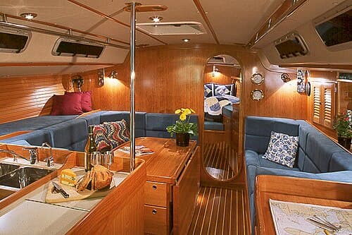 Sailing yacht interior with fold-down table in main salon, galley (kitchen) on left, and navigation station on right and forward cabin visible beyond.